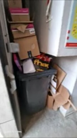 Cartons non aplatis empilés au sol devant un bac d'ordures ménagères saturé